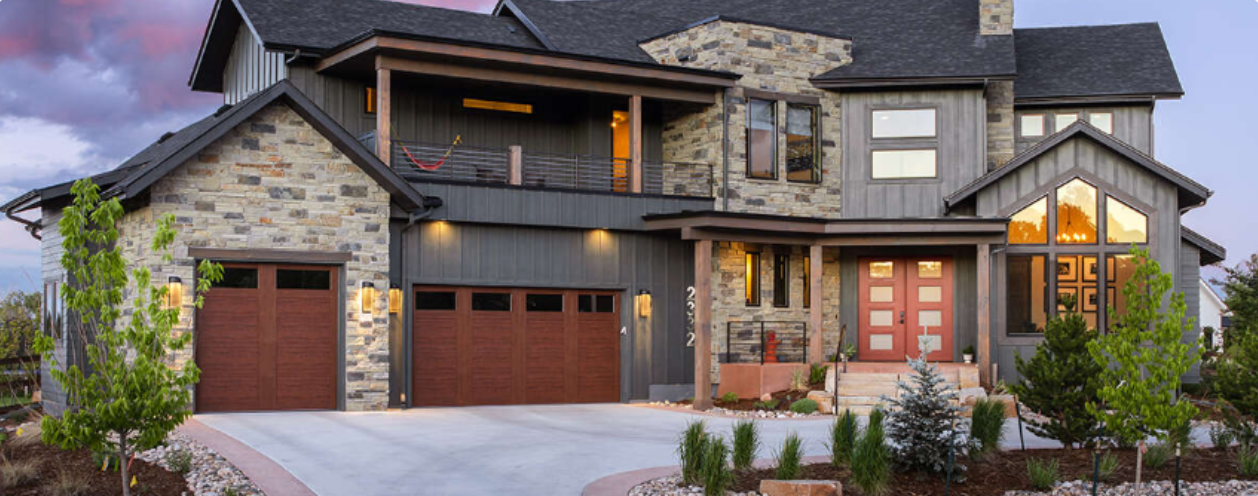 Modern luxury craftsman home with stone and siding exterior, three-car garage, and warm interior lighting at dusk